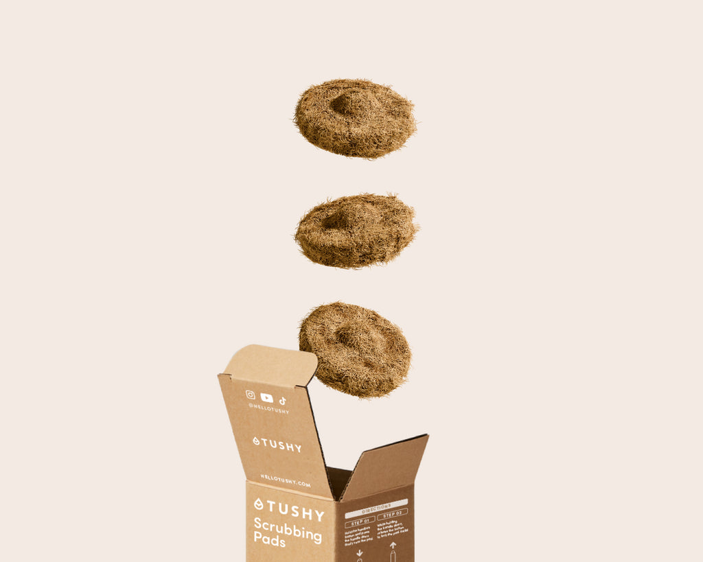 3 Tushy Scrubbing Pads floating above a the box packaging on a beige background