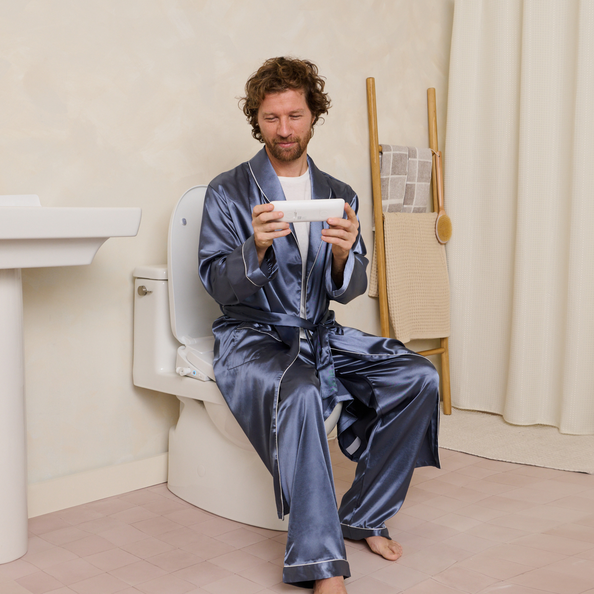 Man in a blue satin pajamas sitting on the toilet looking at his TUSHY Aura remote, in a bathroom setting