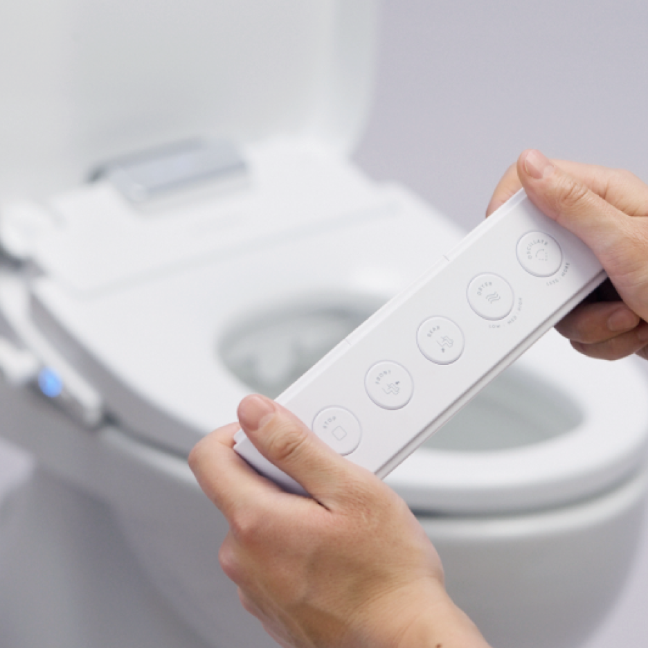Hands holding a TUSHY Aura remote with an out of focus toilet in the background