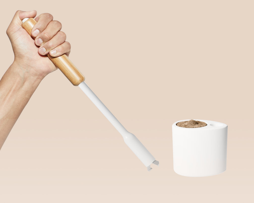 Hand holding a white Tushy toilet brush without a scrubbing pad attached, showin the prongs used to grip the pad. Next to the holder, including a scrubbing pad peeking from the top, on a beige background