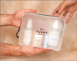 Hands holding an opaque travel case that reads No2 TUSHY BROOKLYN. Inside are 4 TUSHY scented products