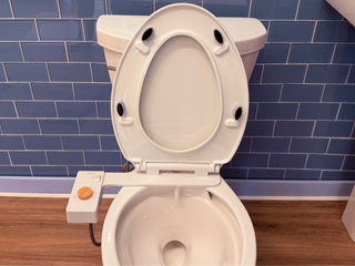 Toilet seat with black bumpers and a control panel on the side, set against a blue tiled wall.