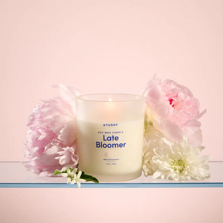 Candle labeled 'Late Bloomer' surrounded by pink and white flowers on a light pink background
