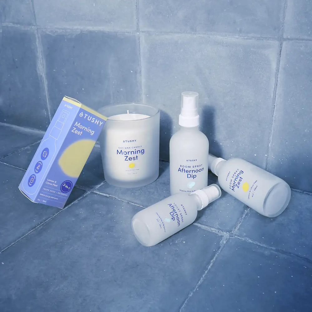 Collection of Tushy products including bottles and a candle on a tiled floor.