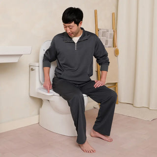 A person sits on a toilet, wearing a gray outfit, with a hand pointing towards the toilet controls.