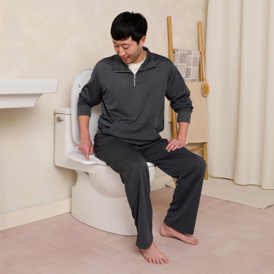 Man in a grey track suit sitting on a toilet, pressing a button on the control panel of a TUSHY Cloud bidet toilet seat.